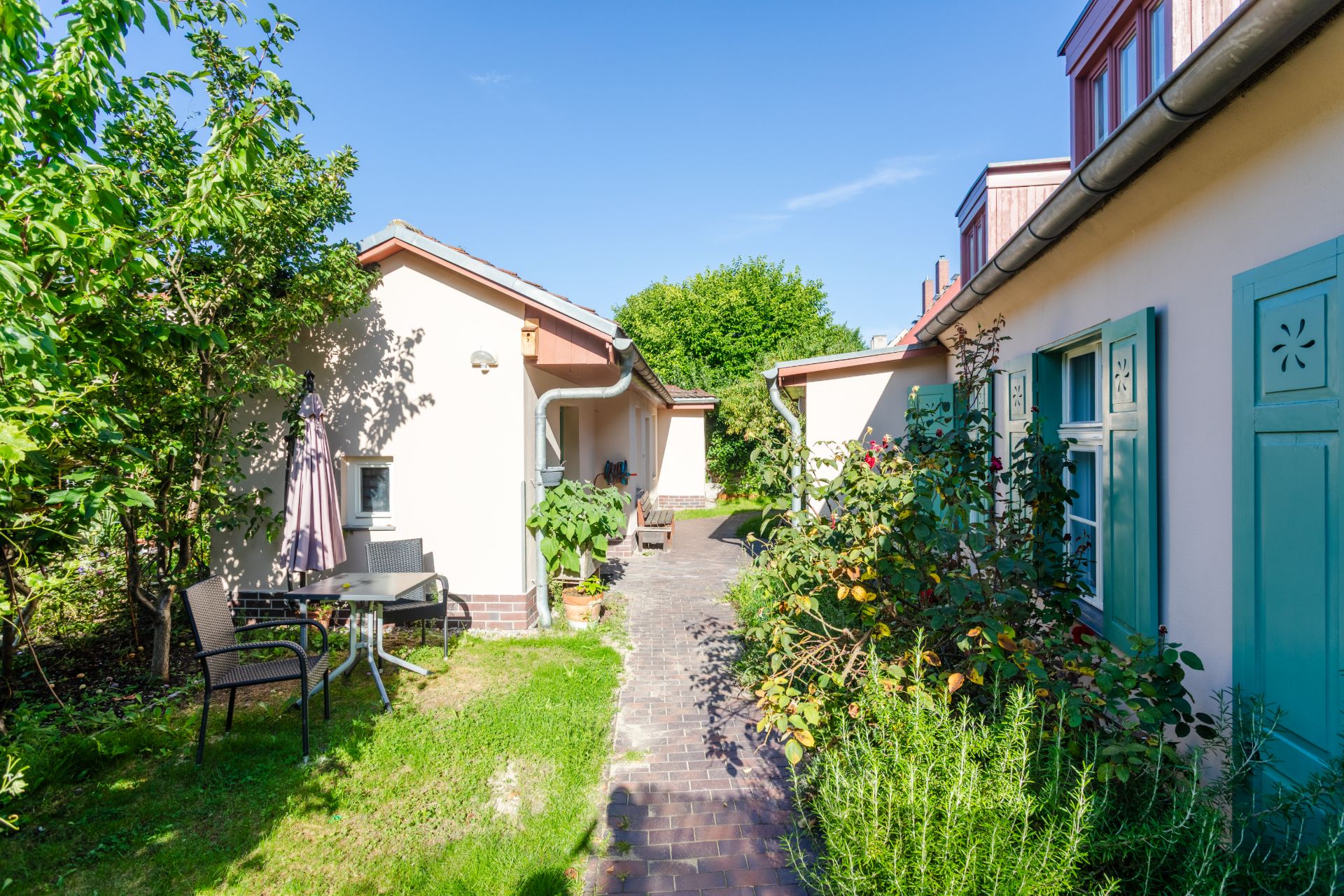 Gepflasterter Weg zwischen zwei Wohnhäusern mit Gartenmöbeln und Pflanzen.
