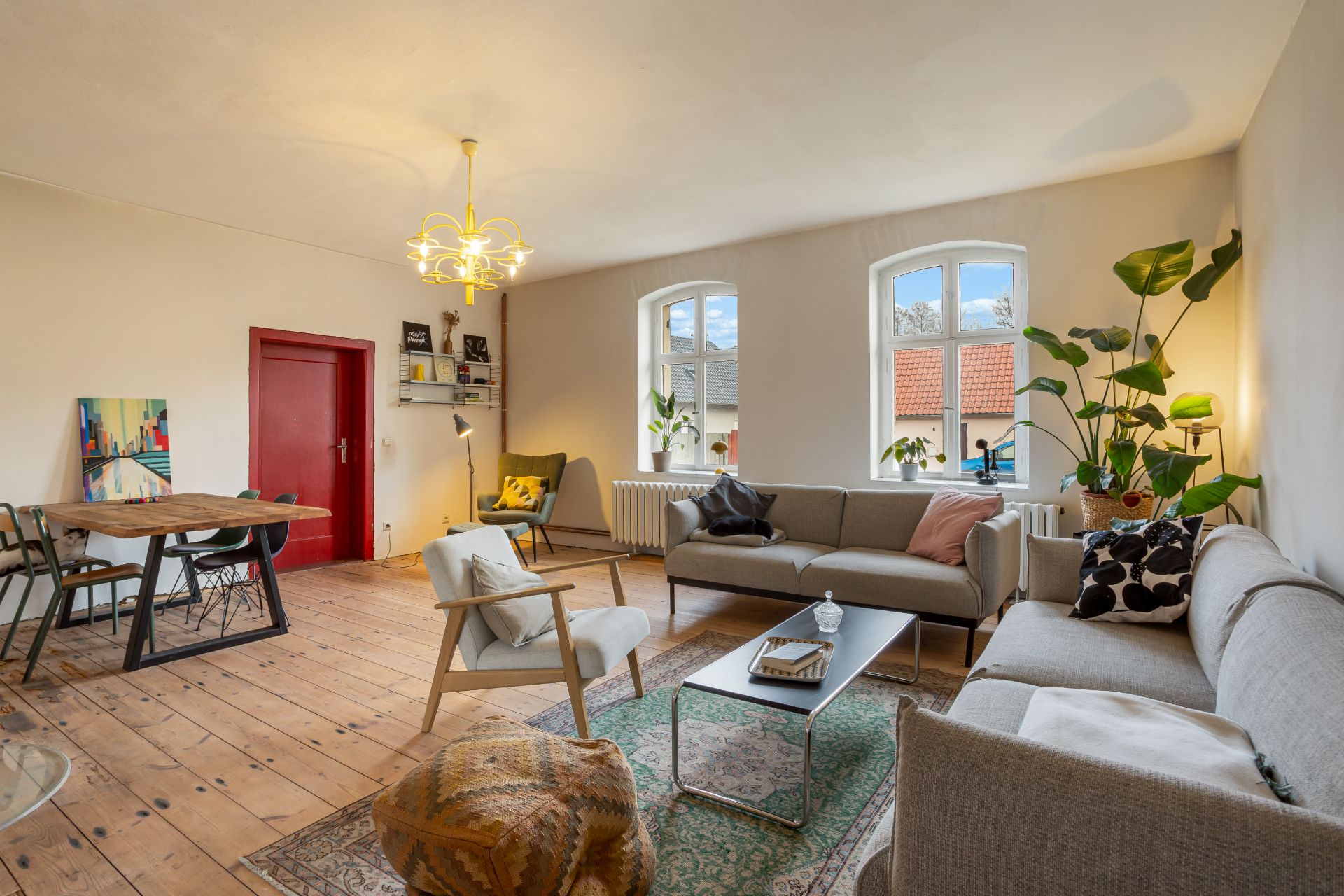 Helles Wohnzimmer mit Holzboden, Sofas, Esstisch, Pflanzen und großen Fenstern.