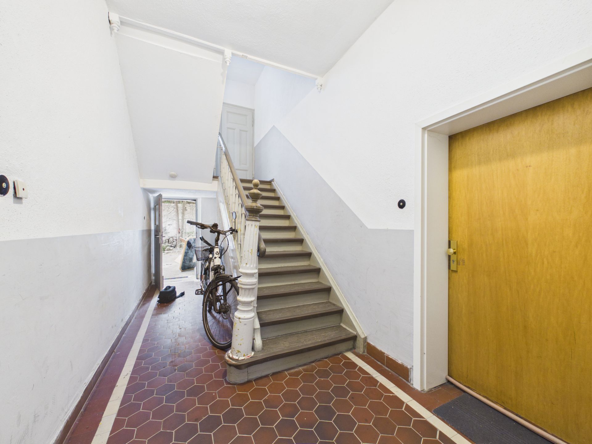 Treppenhaus mit Fahrrad, braunen Bodenfliesen und gelber Tür, Tageslicht von außen.