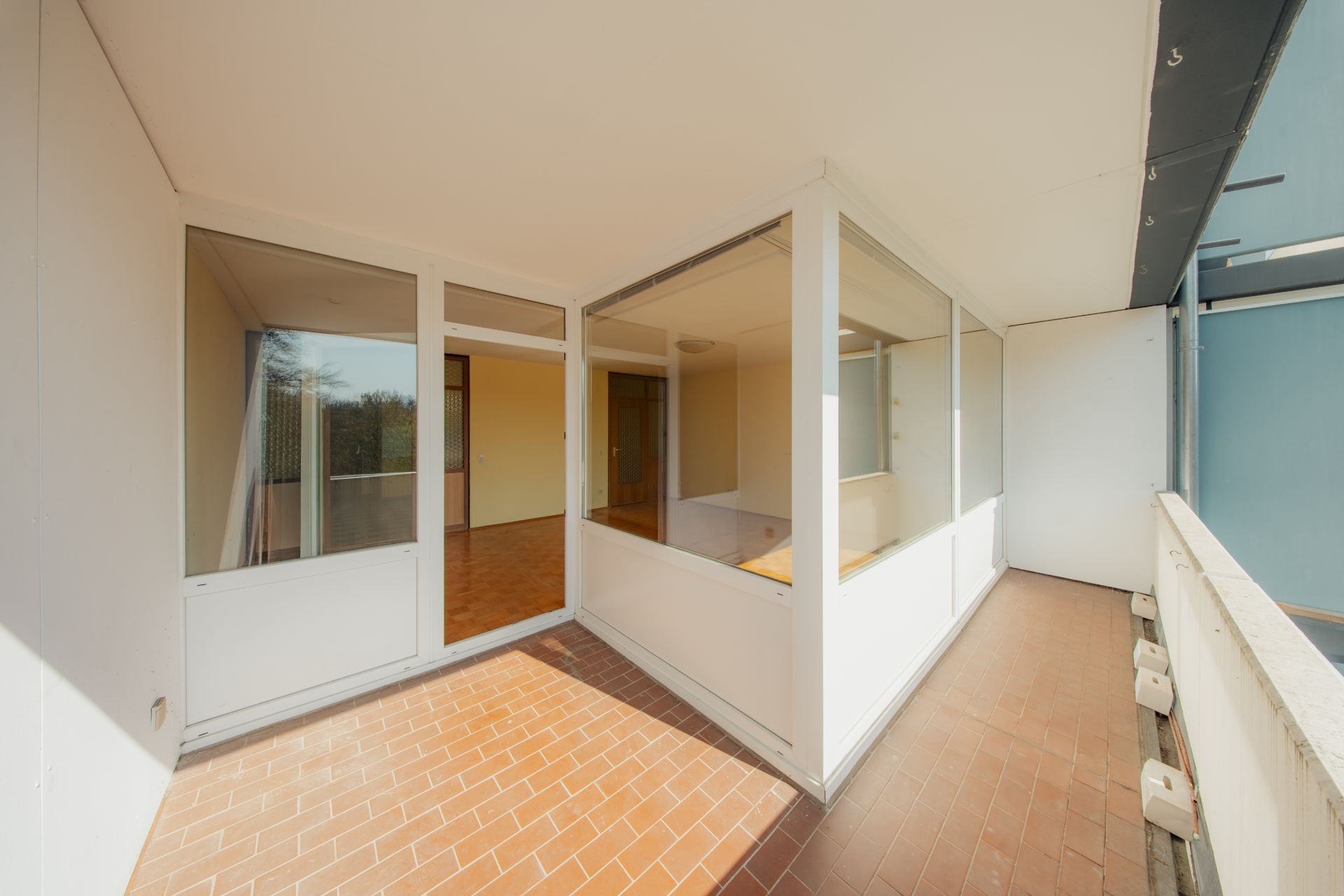 Überdachter Balkon mit Glaswänden und braunem Fliesenboden, angrenzend an Innenräume.