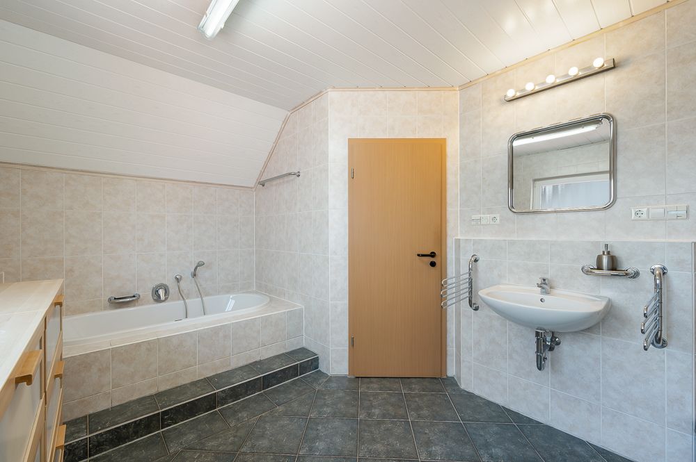 Badezimmer mit Badewanne, Waschbecken, Spiegel und Holztür, gefliest und mit schräger Decke.