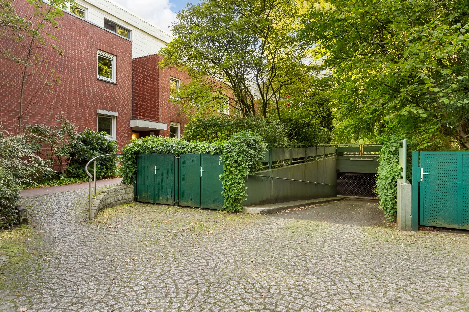 Eingang zu einer Tiefgarage mit grünem Tor neben einem roten Wohngebäude und vielen Bäumen.