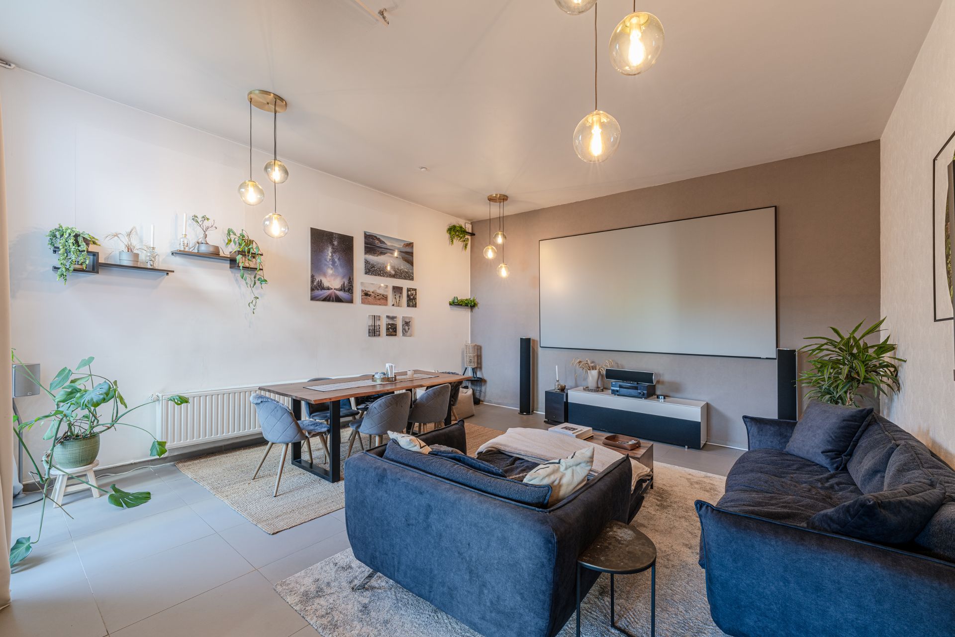 Modernes Wohnzimmer mit blauen Sofas, Esstisch, Pflanzen und großer Leinwand an der Wand.