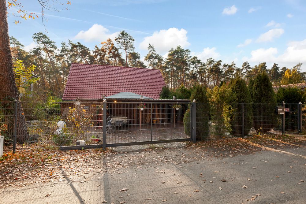 Haus mit rotem Dach hinter einem Metallzaun, umgeben von Bäumen und Sträuchern.