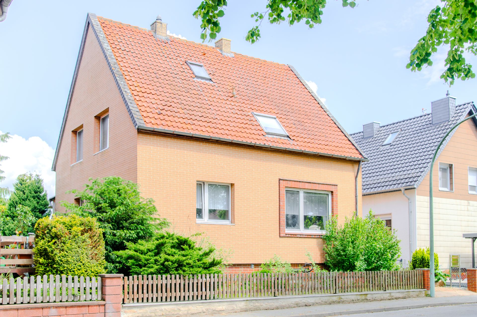 Zweistöckiges Haus mit rotem Satteldach, Garten und Holzzaun an einer Straße.