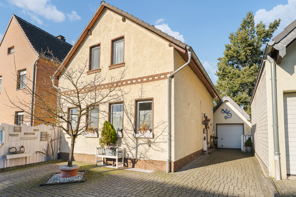 Zweistöckiges Haus mit hellem Putz, Innenhof, kleinem Baum und Garage im Hintergrund.