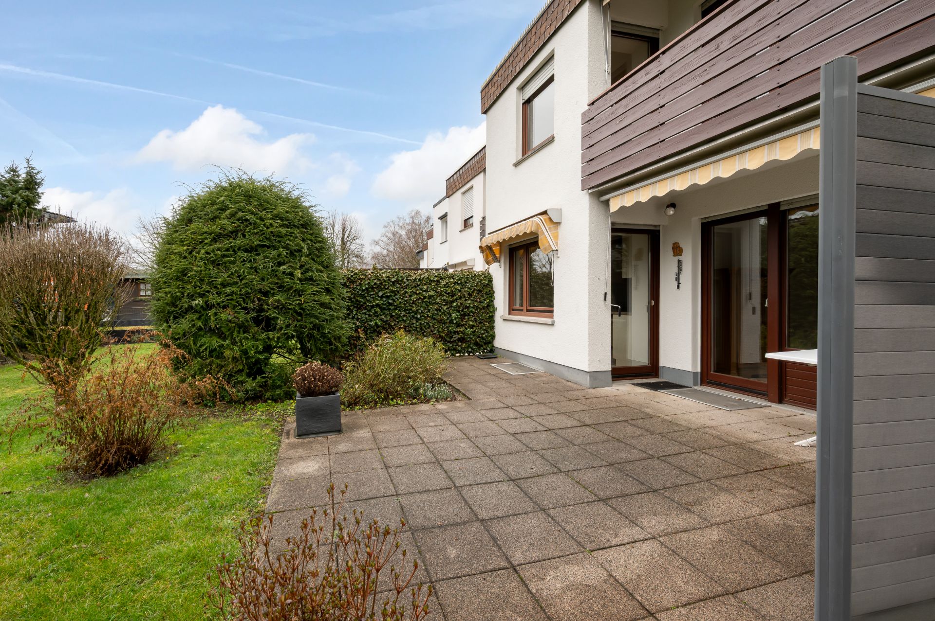 Gepflasterte Terrasse mit Markise und Zugang zum Garten vor einem modernen Wohnhaus.