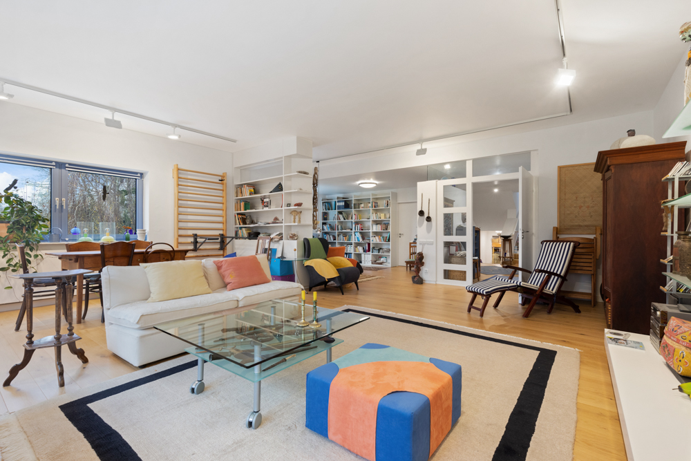 Helles Wohnzimmer mit Sofa, Glastisch, buntem Hocker, Bücherregal und großen Fenstern.