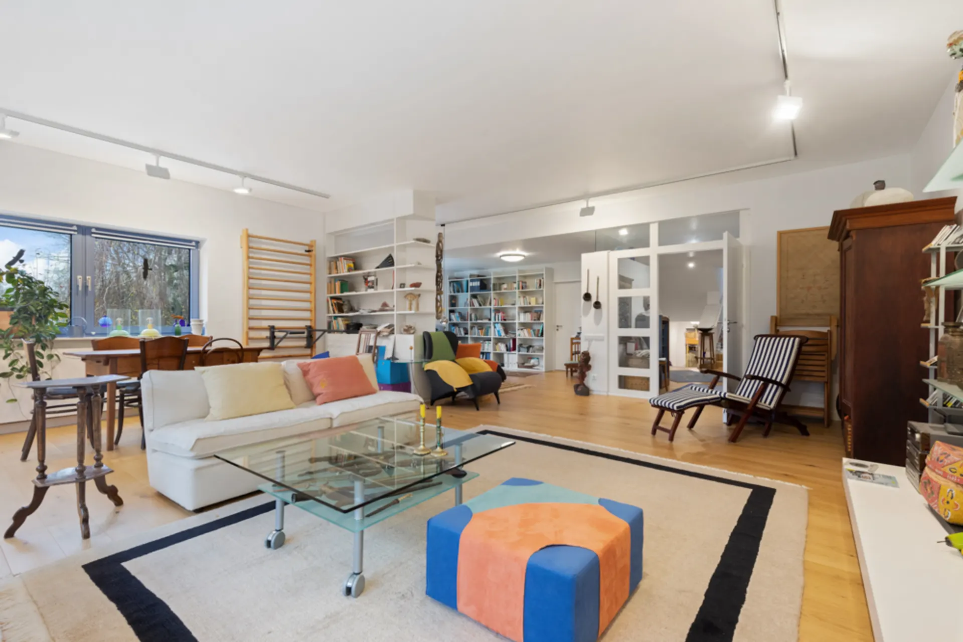 Helles Wohnzimmer mit Sofa, Glastisch, buntem Hocker, Bücherregal und großen Fenstern.