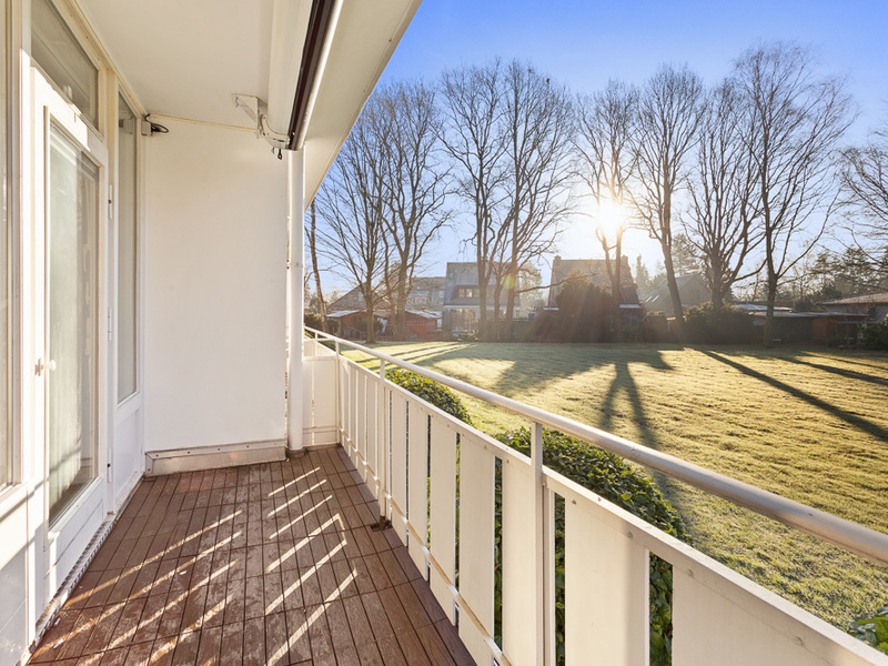 Sonniger Balkon mit Holzboden und Blick auf einen Garten mit Bäumen und Rasen.