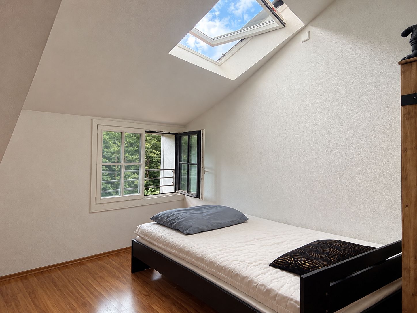 Helles Schlafzimmer mit Einzelbett, Dachfenster und offenem Fenster, Holzboden.