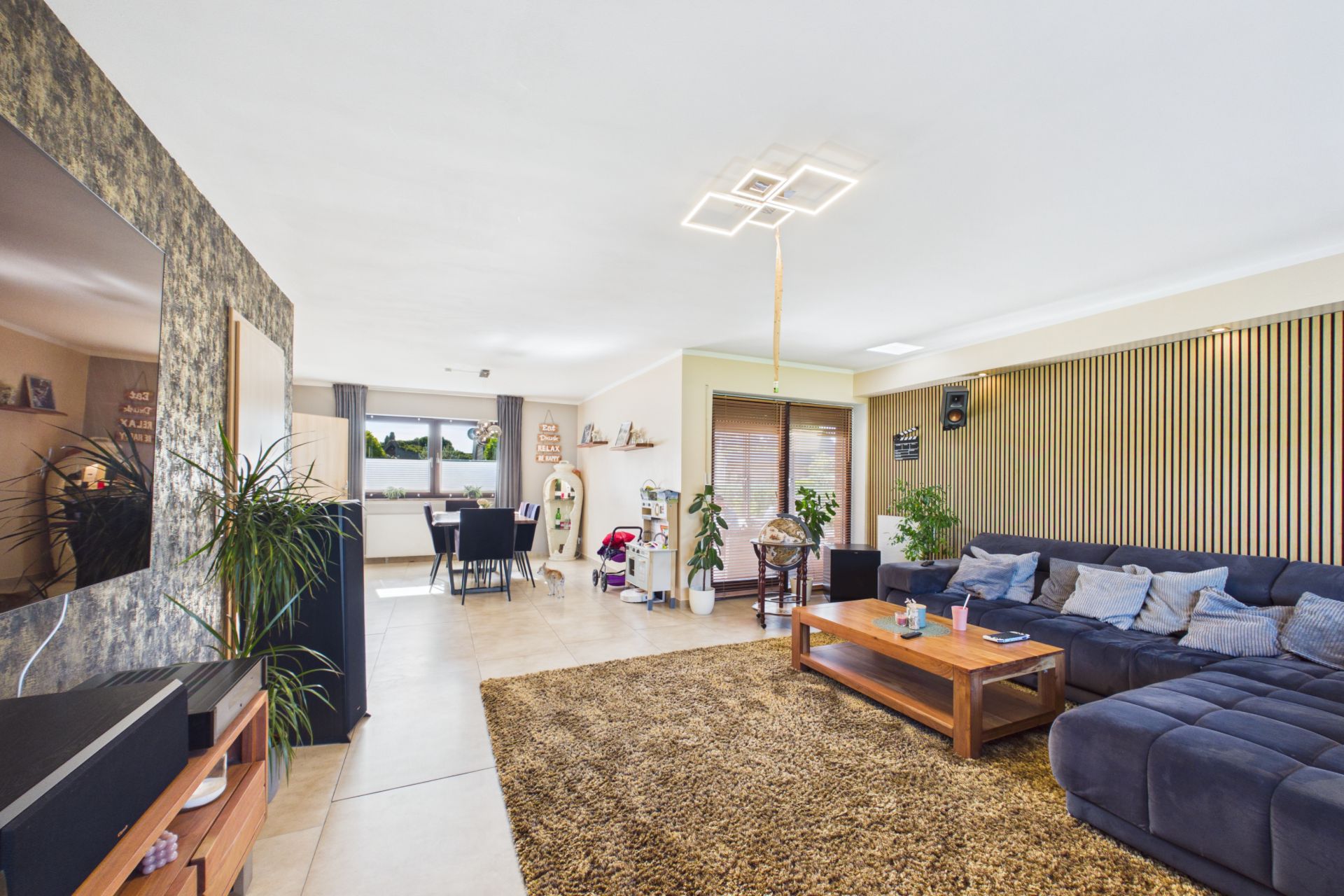 Helles Wohnzimmer mit Ecksofa, Teppich, Couchtisch und Essbereich im Hintergrund.