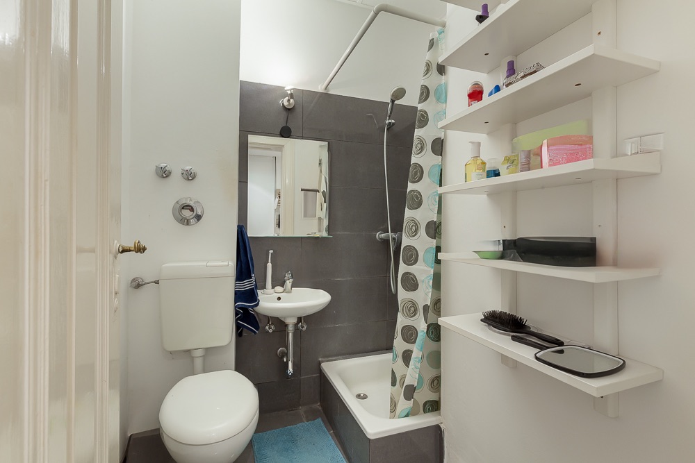 Kleines Badezimmer mit Toilette, Waschbecken, Dusche mit Duschvorhang und weißen Wandregalen.