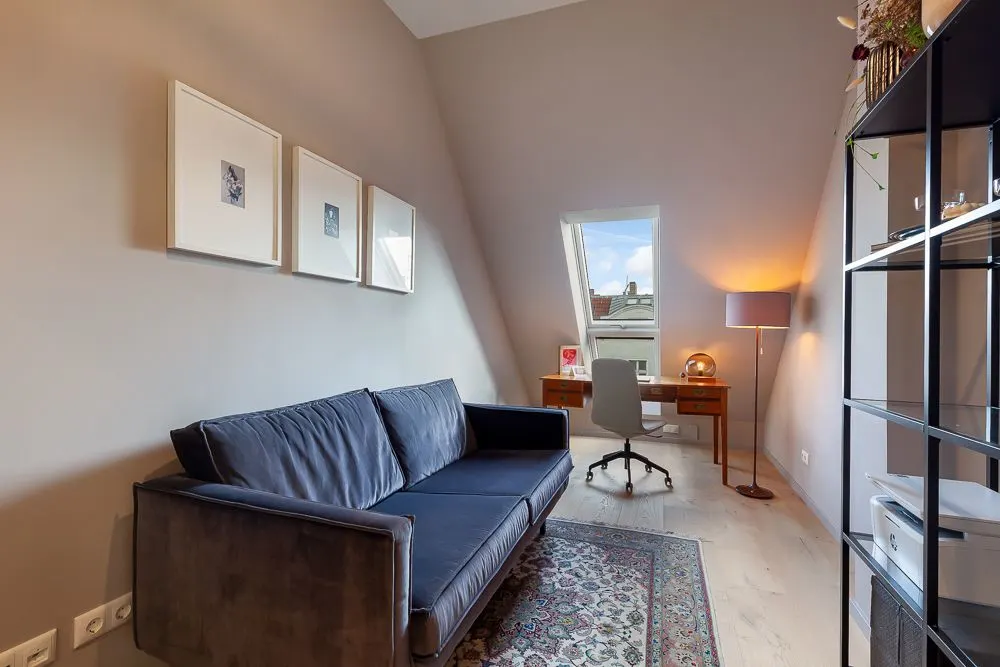 Helles Arbeitszimmer mit Sofa, Schreibtisch am Fenster und Regal an der Wand.