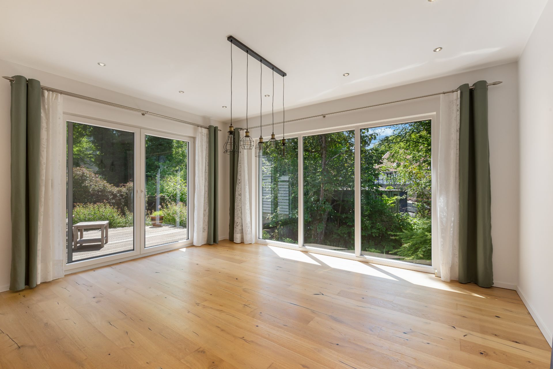 Heller Raum mit Holzboden, großen Fenstern und Blick auf eine Terrasse und Garten.