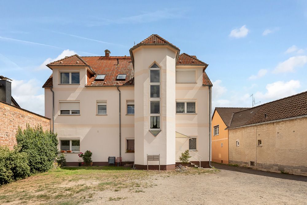 Dreistöckiges Wohnhaus mit rotem Dach und markantem Erker in der Mitte, umgeben von anderen Gebäuden.