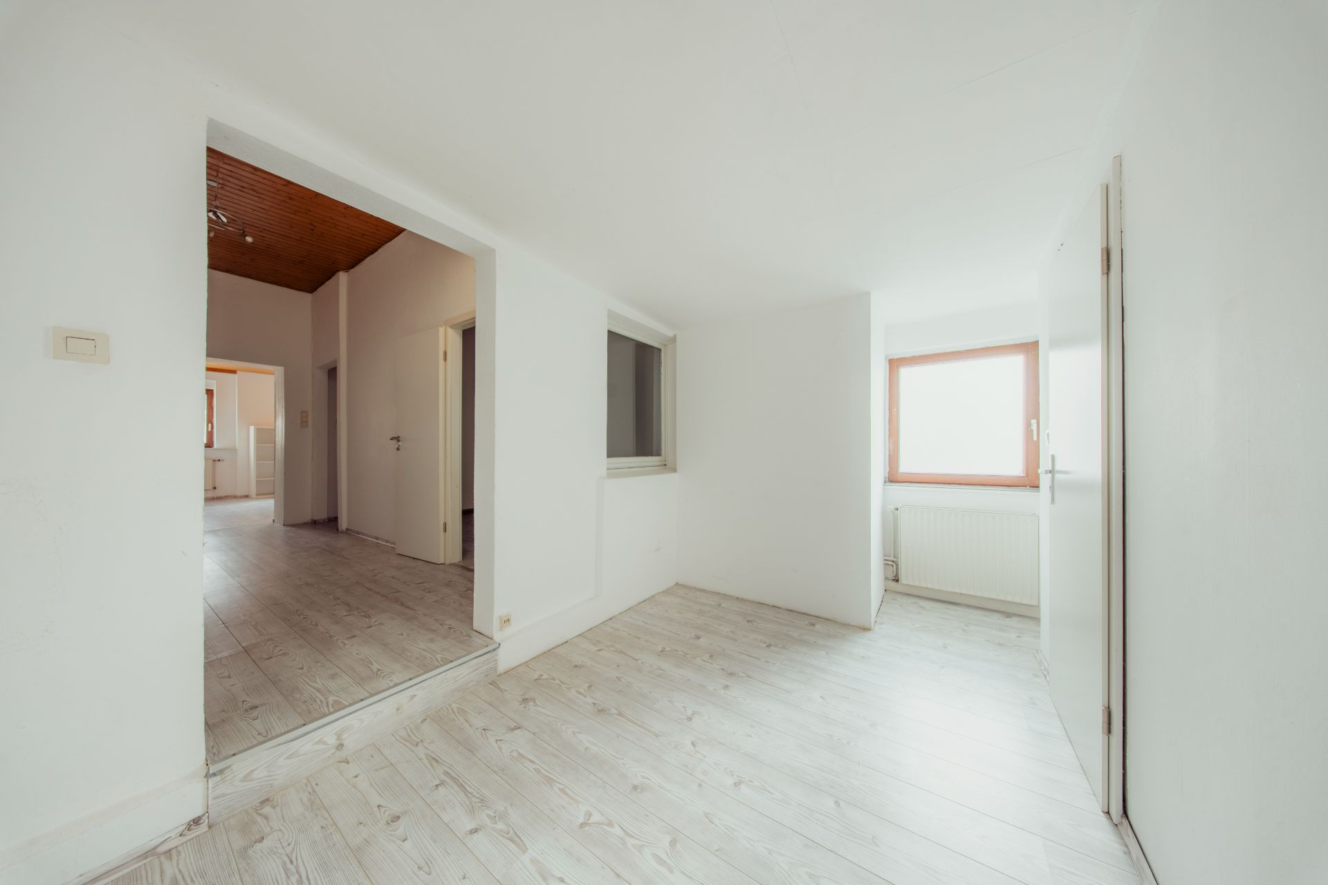 Helles, leeres Zimmer mit Holzfußboden, Fenster und Türen, weiße Wände.