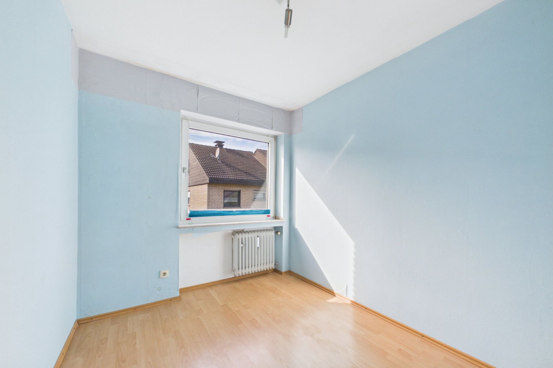 Helles, leeres Zimmer mit hellblauen Wänden, Holzfußboden und großem Fenster mit Blick nach draußen.
