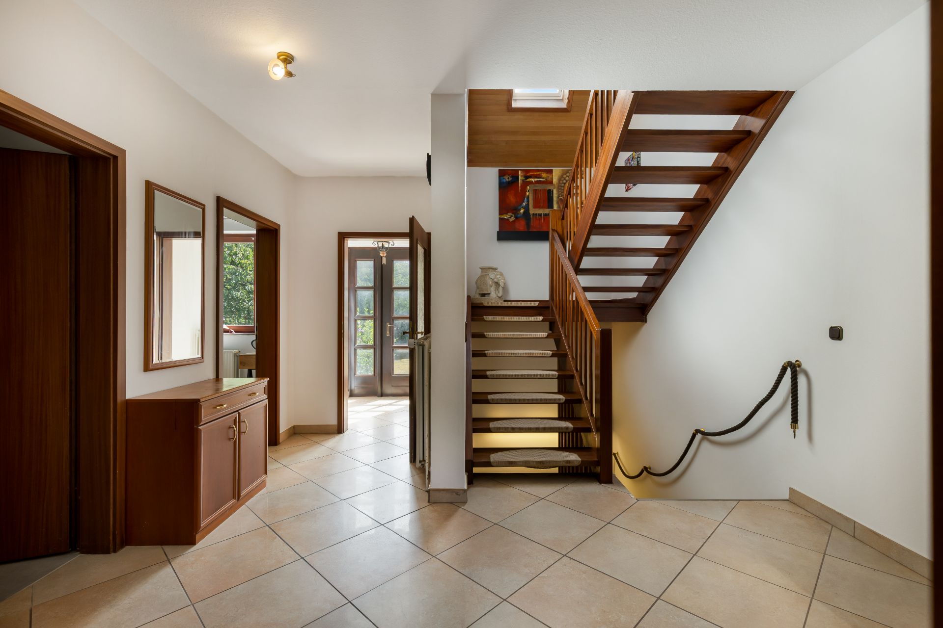 Heller Flur mit Holztreppe, Kommode, Spiegeln und Fliesenboden, Tür führt nach draußen.