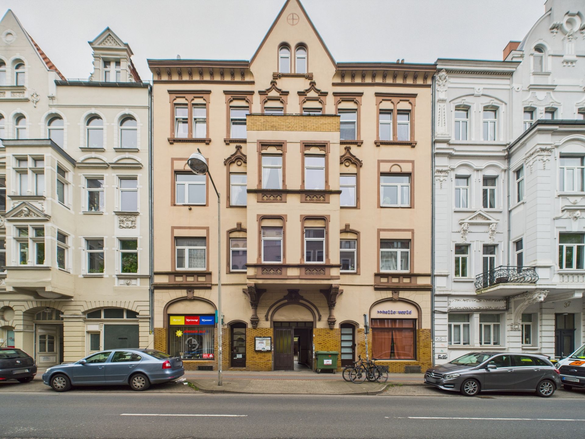 Mehrstöckiges Wohnhaus mit Verzierungen und Geschäften im Erdgeschoss, Autos und Fahrräder davor.