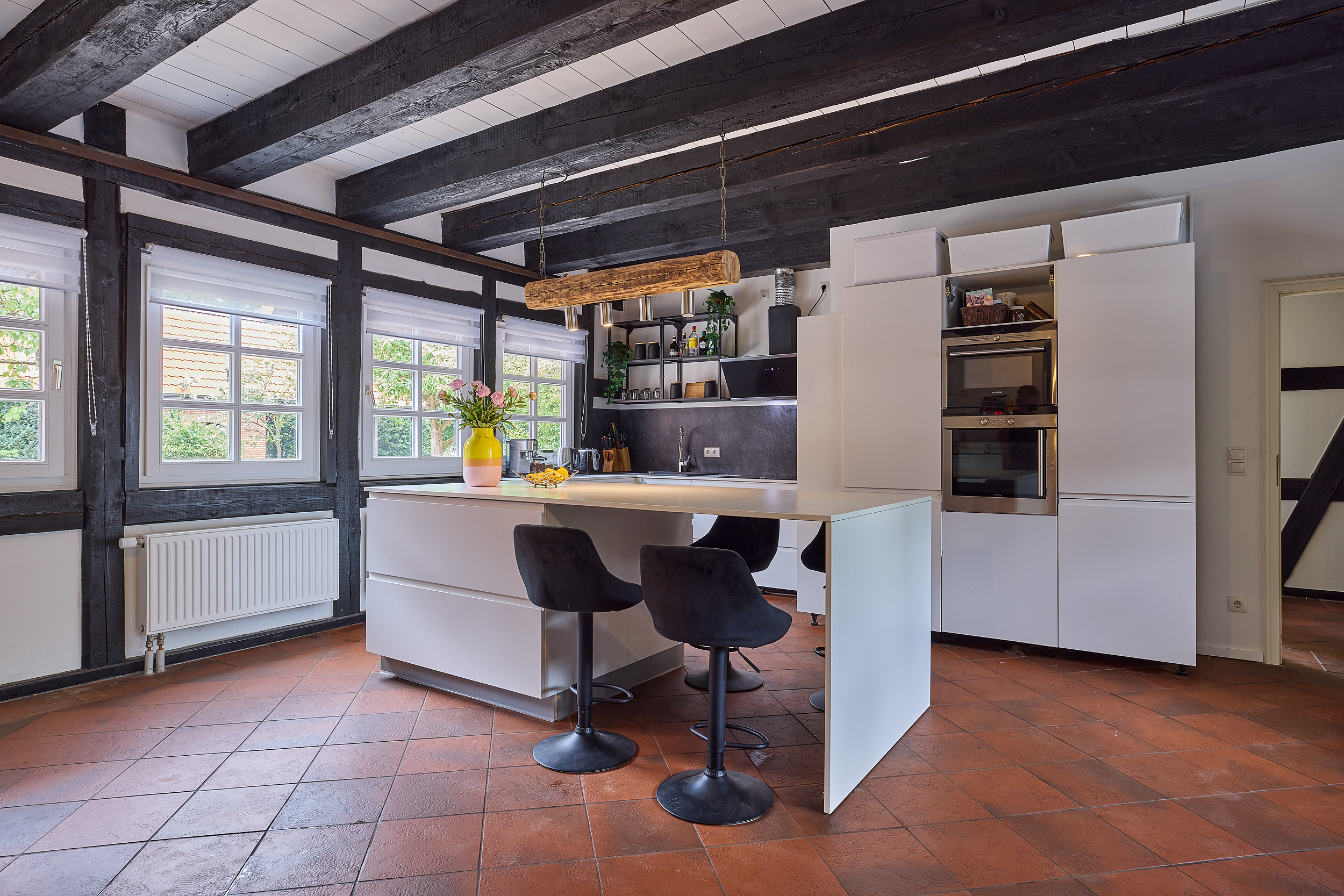Moderne Küche mit weißer Insel, schwarzen Stühlen und großen Fenstern mit sichtbaren Holzbalken.