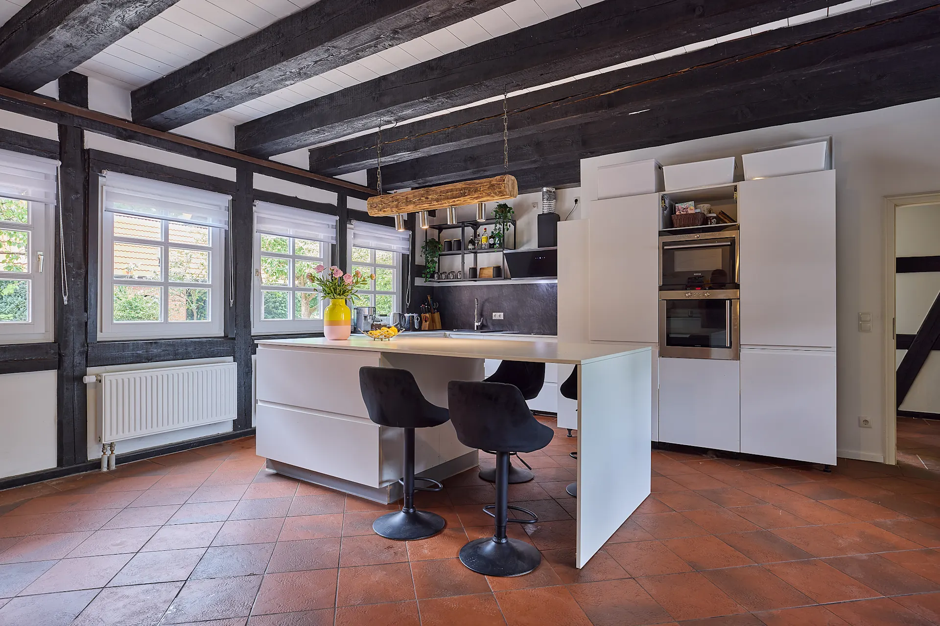 Moderne Küche mit weißer Insel, schwarzen Stühlen und großen Fenstern mit sichtbaren Holzbalken.