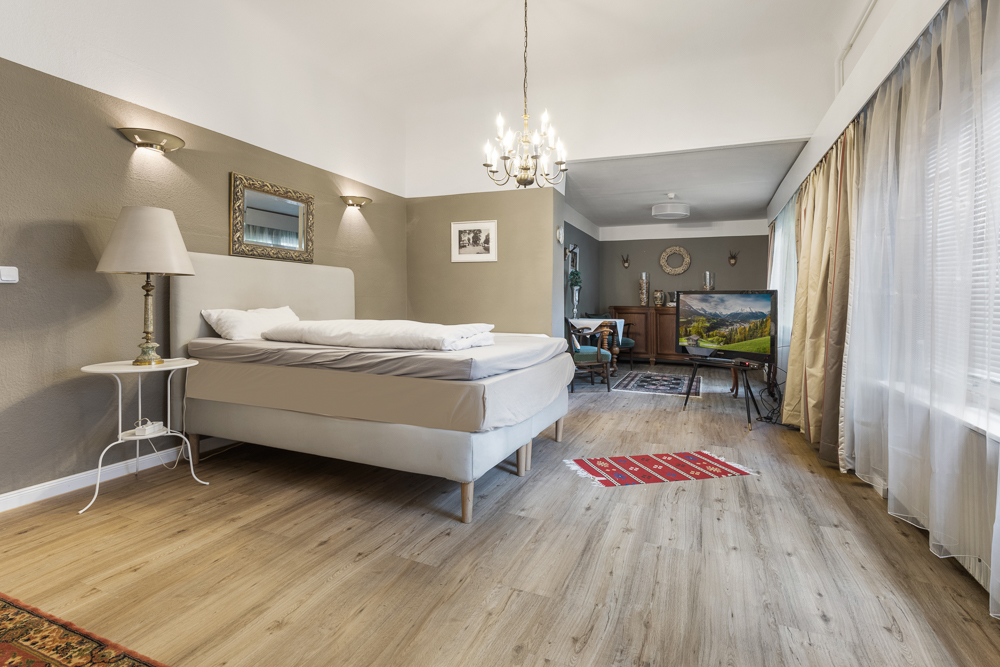 Helles Schlafzimmer mit großem Bett, Nachttisch, Stehlampe, Fernseher und großen Fenstern mit Vorhängen.