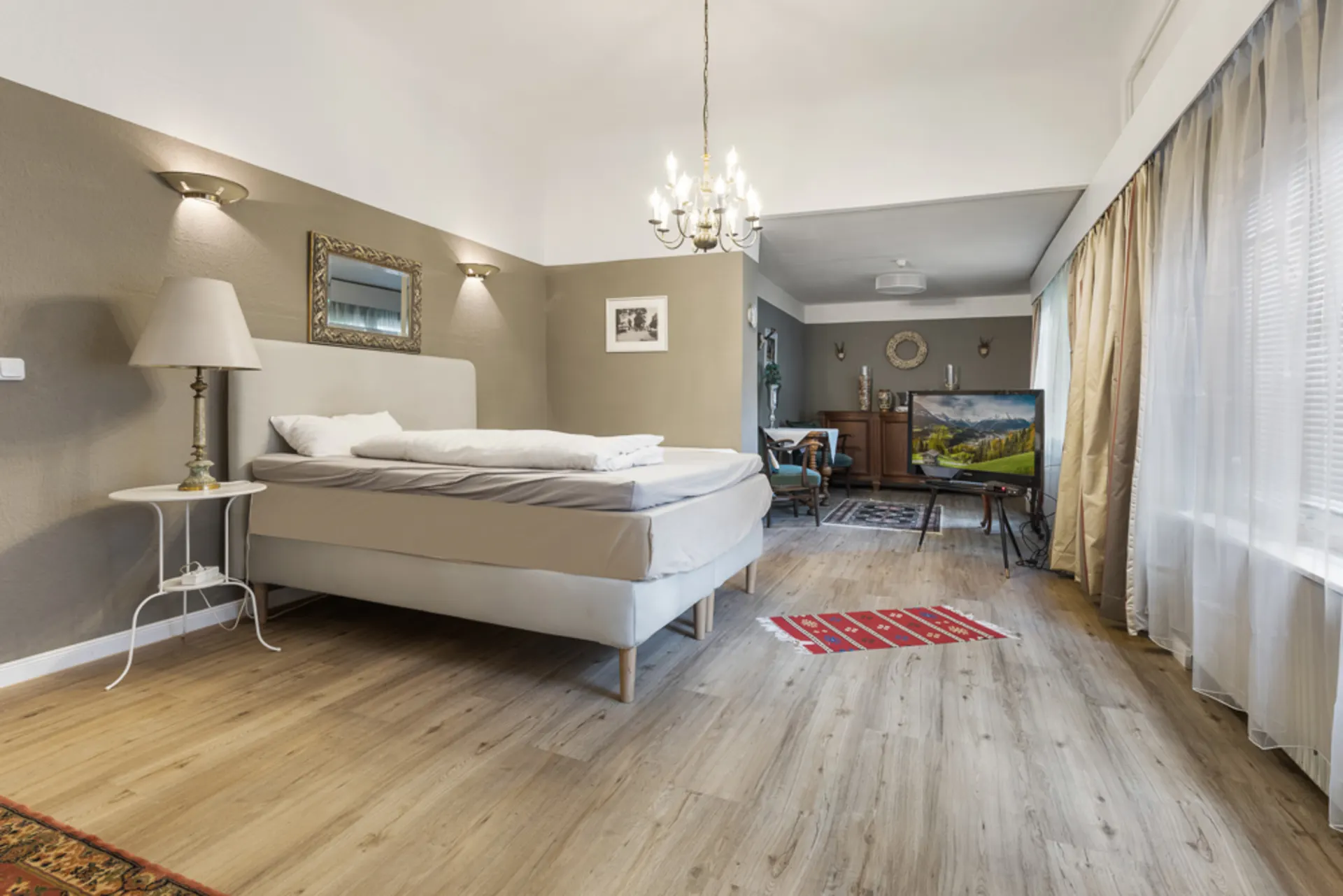 Helles Schlafzimmer mit großem Bett, Nachttisch, Stehlampe, Fernseher und großen Fenstern mit Vorhängen.