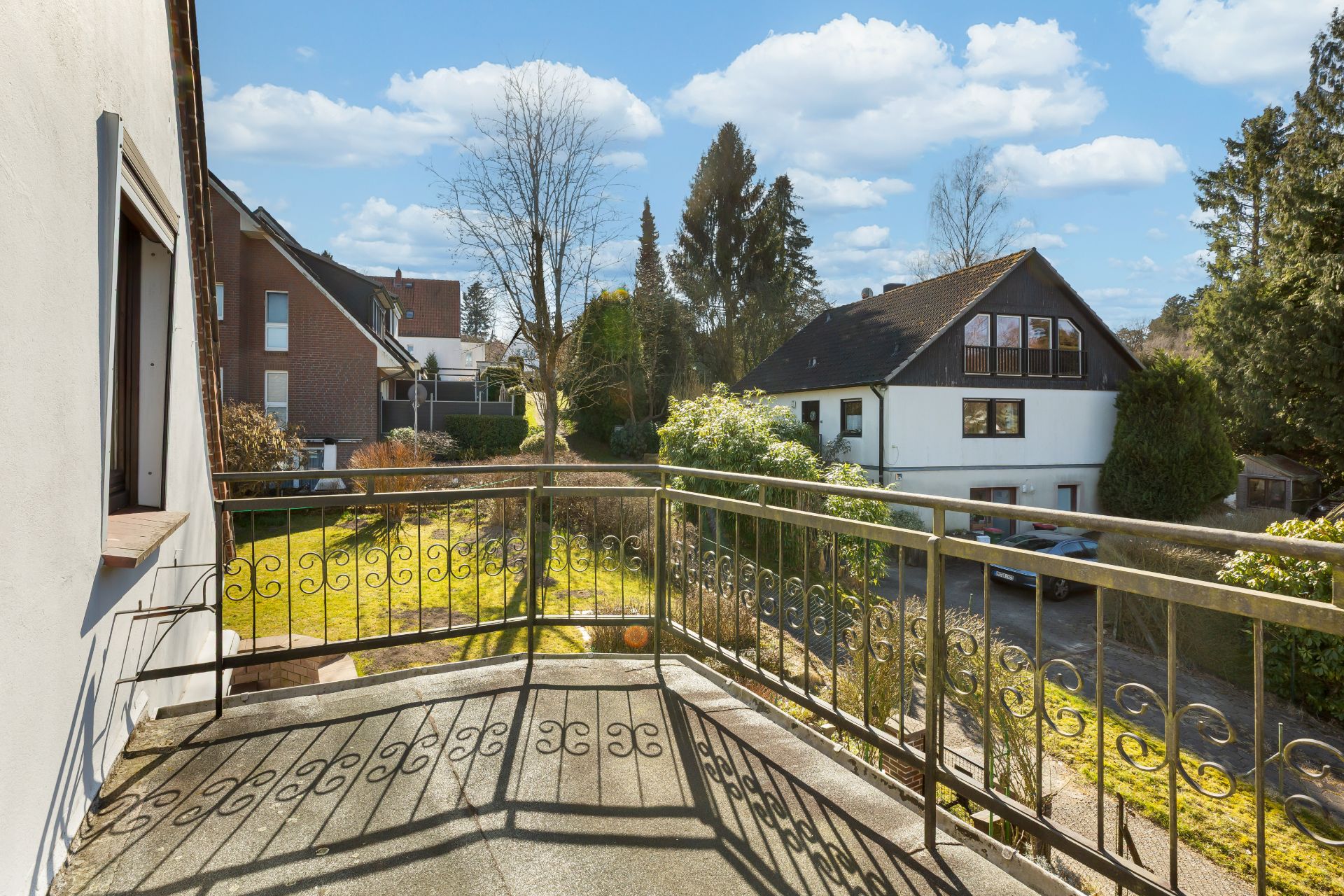 Balkon mit Metallgeländer, Blick auf Garten und Nachbarhäuser an einem sonnigen Tag.