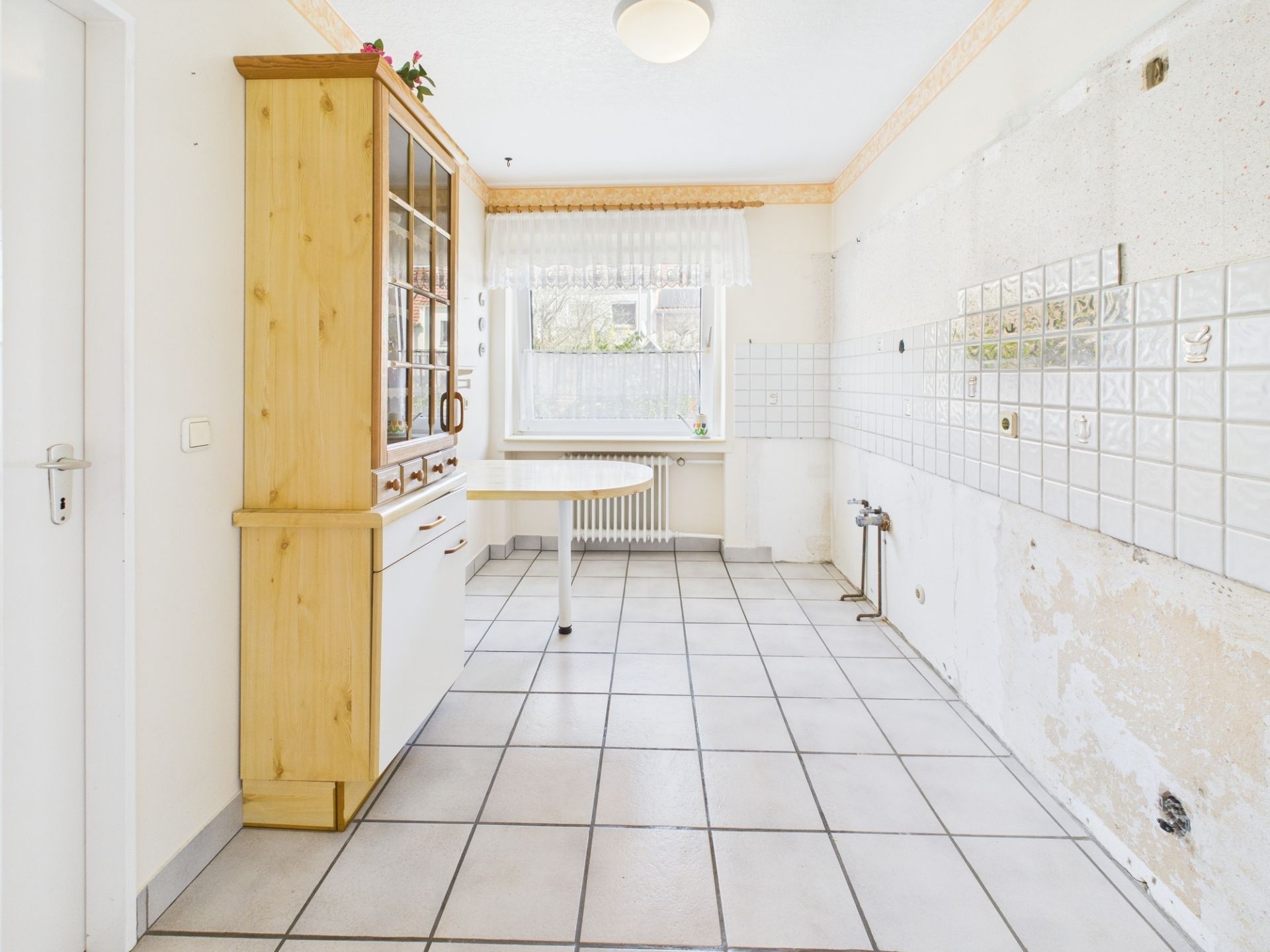 Leere Küche mit Fliesenboden, Holzschrank und einem Fenster mit Gardine.