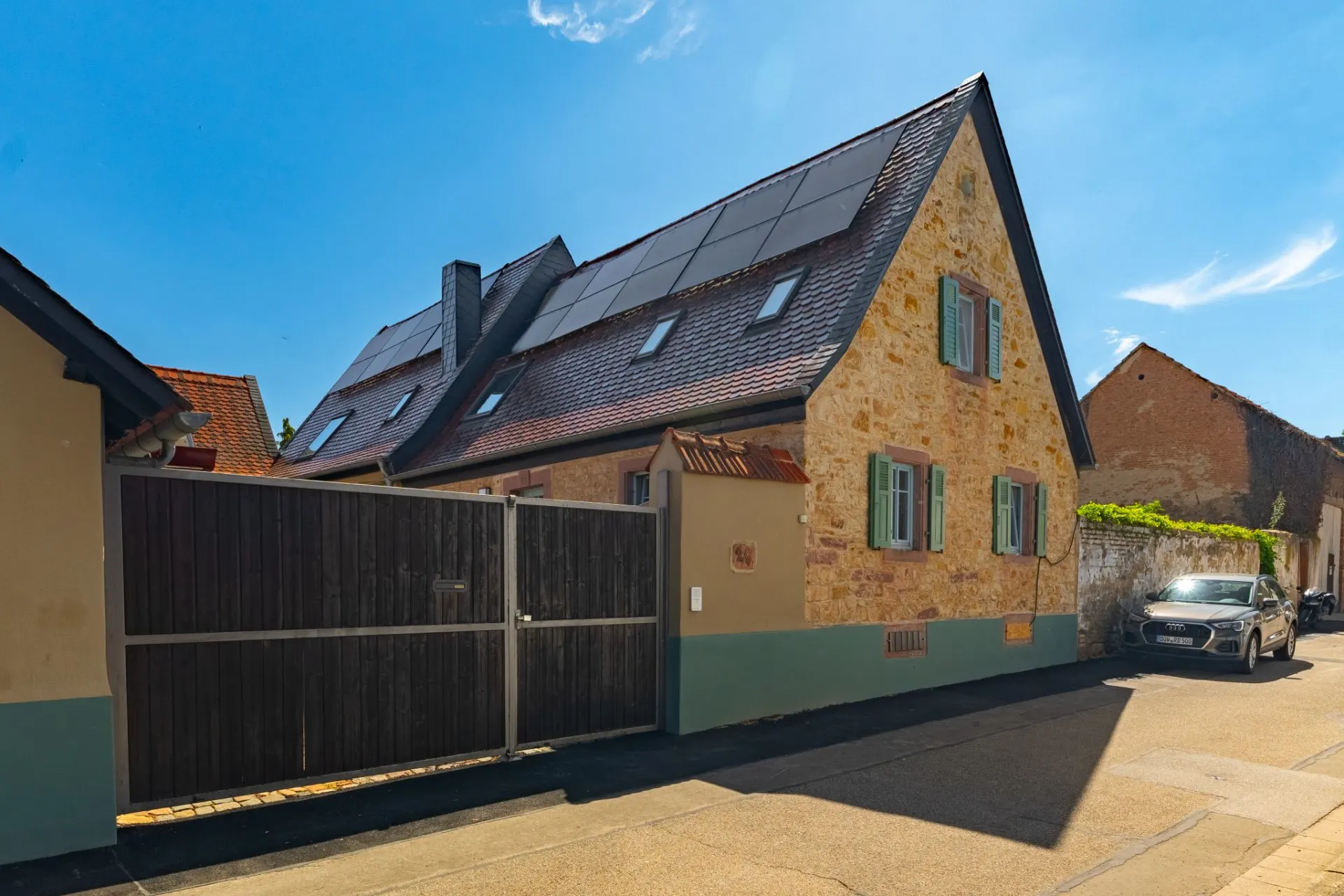 Steinhaus mit Solarpanels auf dem Dach, grüne Fensterläden und Holztor an einer ruhigen Straße.