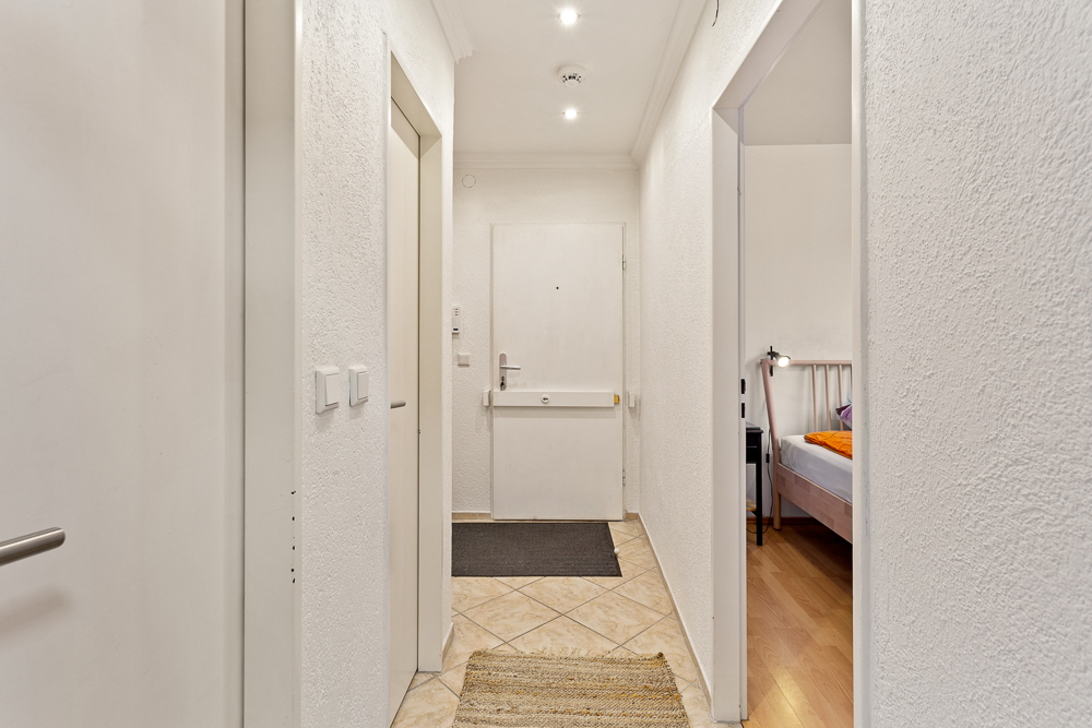 Heller Flur mit Fliesenboden, weißer Haustür und Blick in ein Schlafzimmer.