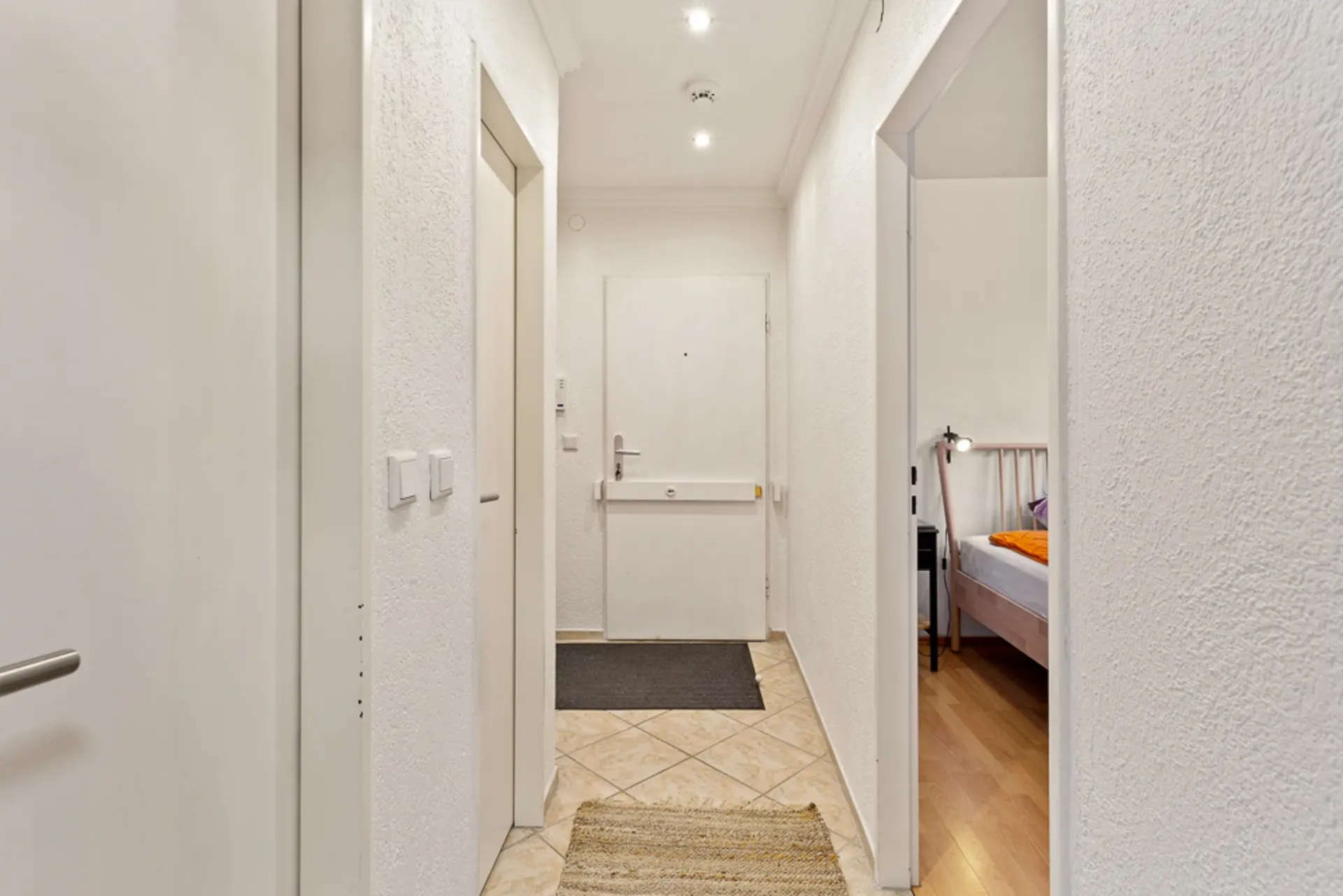 Heller Flur mit Fliesenboden, weißer Haustür und Blick in ein Schlafzimmer.