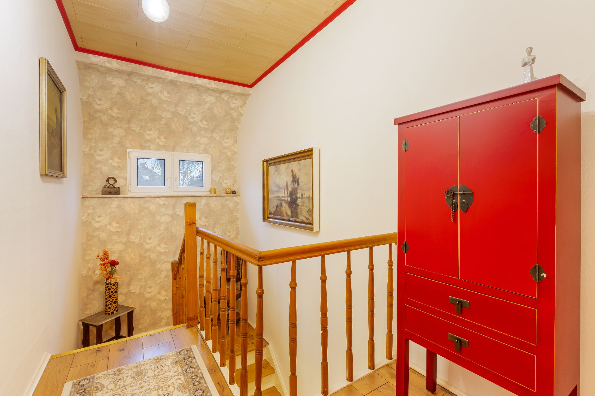 Heller Flur mit Holztreppe, rotem Schrank, Teppich und kleinen Fenstern.