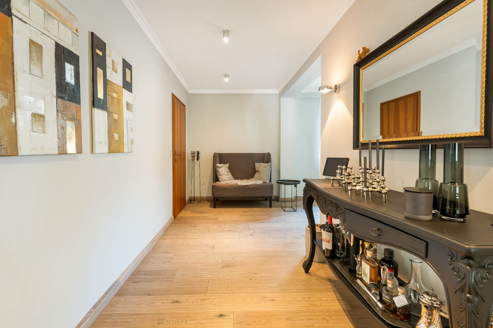 Heller Flur mit Holzfußboden, Gemälden an der Wand, Sofa und dunklem Sideboard mit Spiegel.