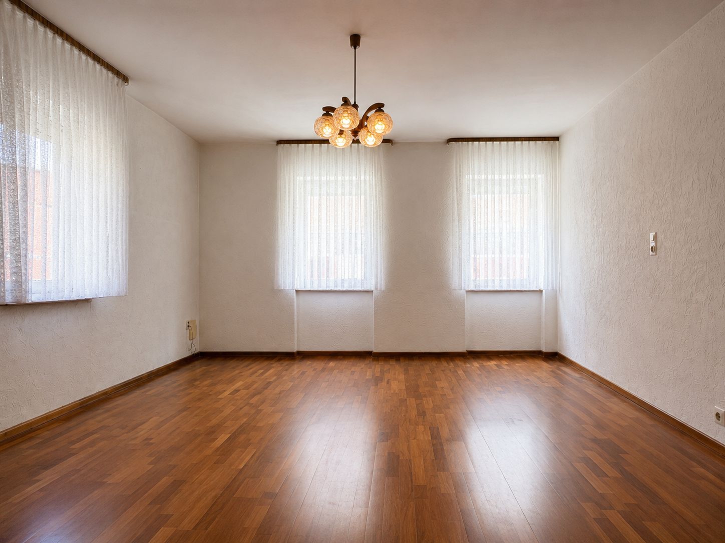 Leeres Zimmer mit Holzfußboden, drei Fenstern mit Gardinen und Deckenleuchte.