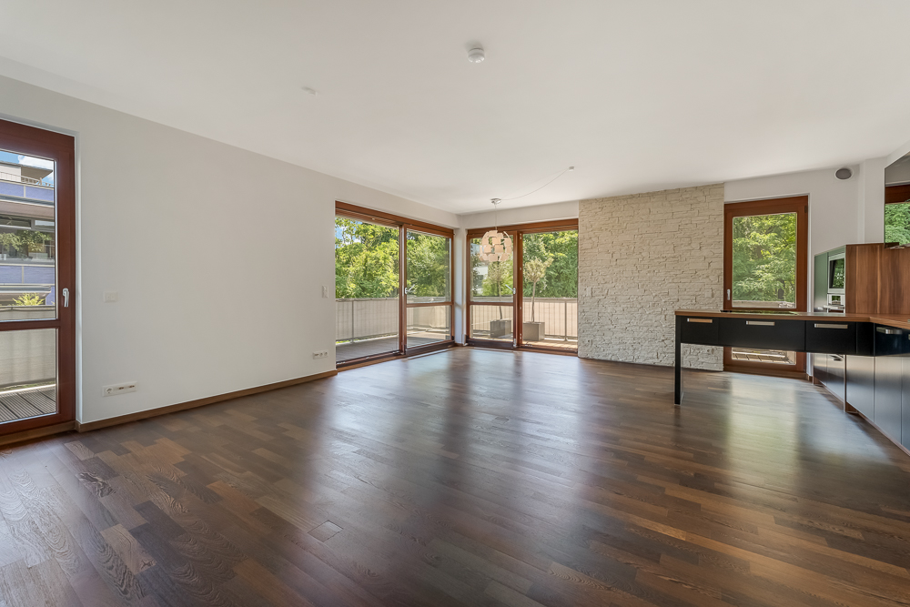 Helles, leeres Wohnzimmer mit dunklem Holzboden, großen Fenstern und offener Küche.