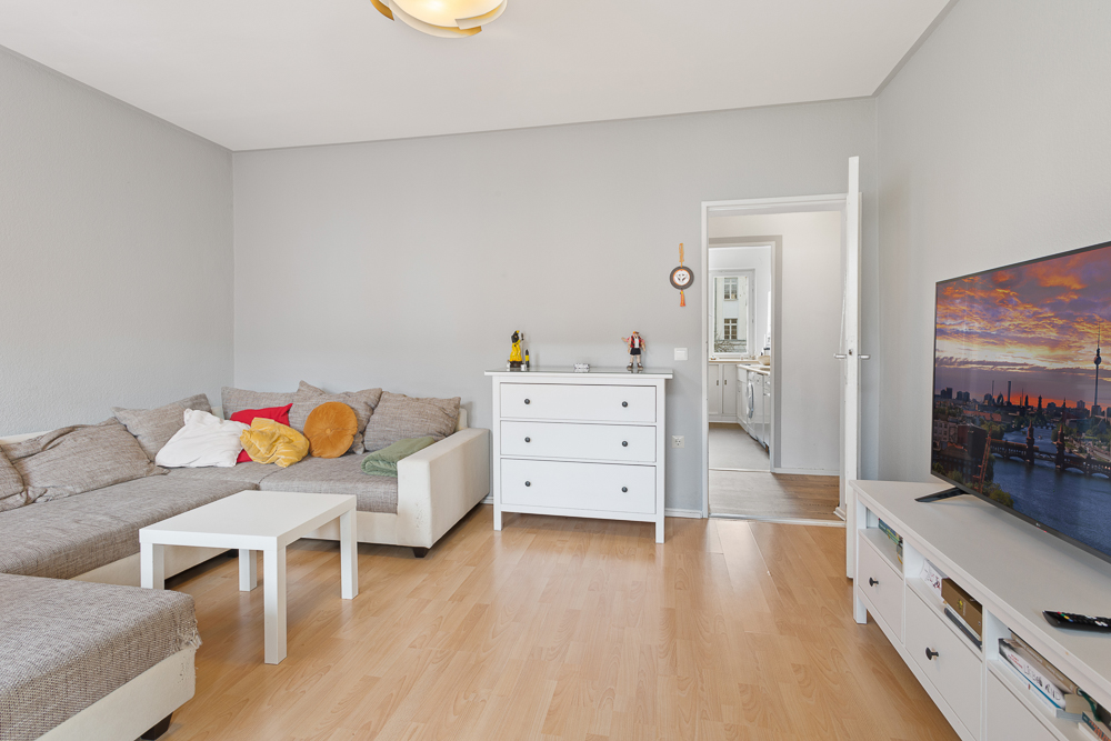 Wohnzimmer mit Ecksofa, weißem Couchtisch, Kommode und Fernseher auf hellem Holzboden.