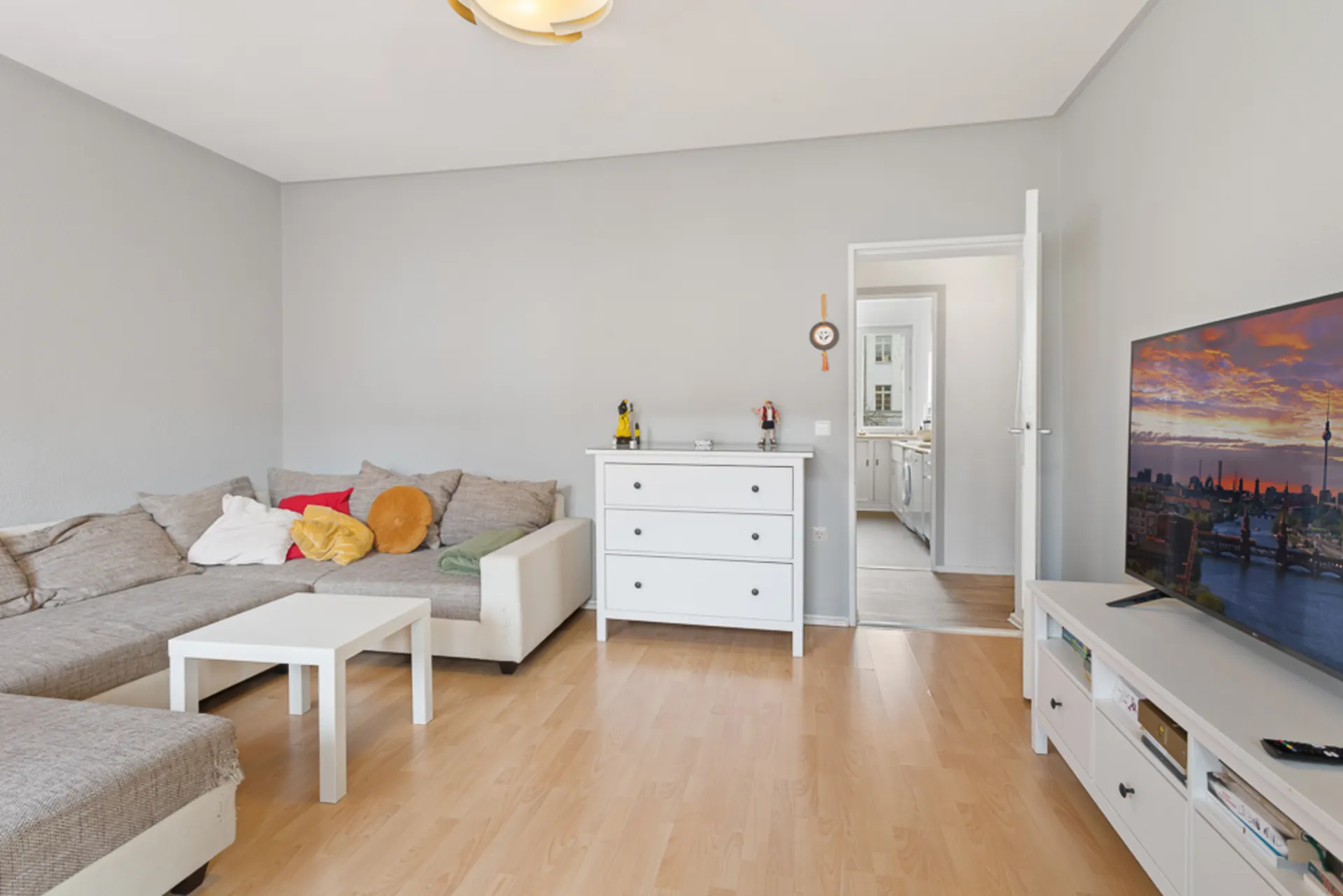 Wohnzimmer mit Ecksofa, weißem Couchtisch, Kommode und Fernseher auf hellem Holzboden.