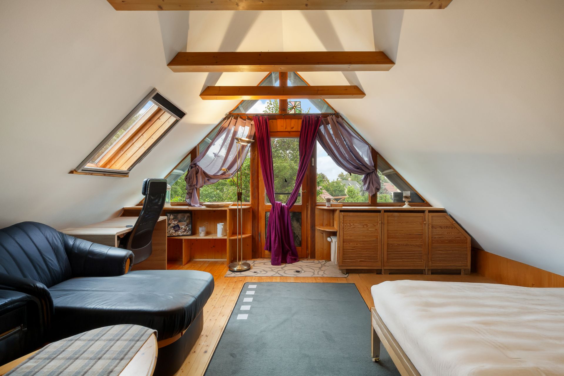 Dachzimmer mit Holzbalken, Sofa, Schreibtisch, Bett und Tür mit lila Vorhängen.