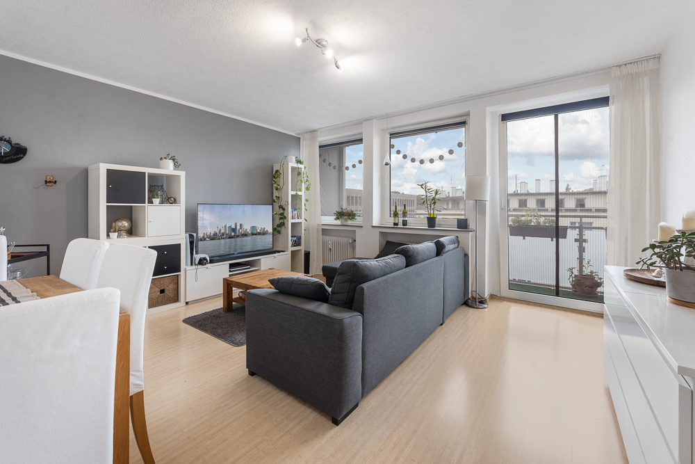 Helles Wohnzimmer mit grauer Couch, Fernseher, Esstisch und großen Fenstern mit Balkonzugang.