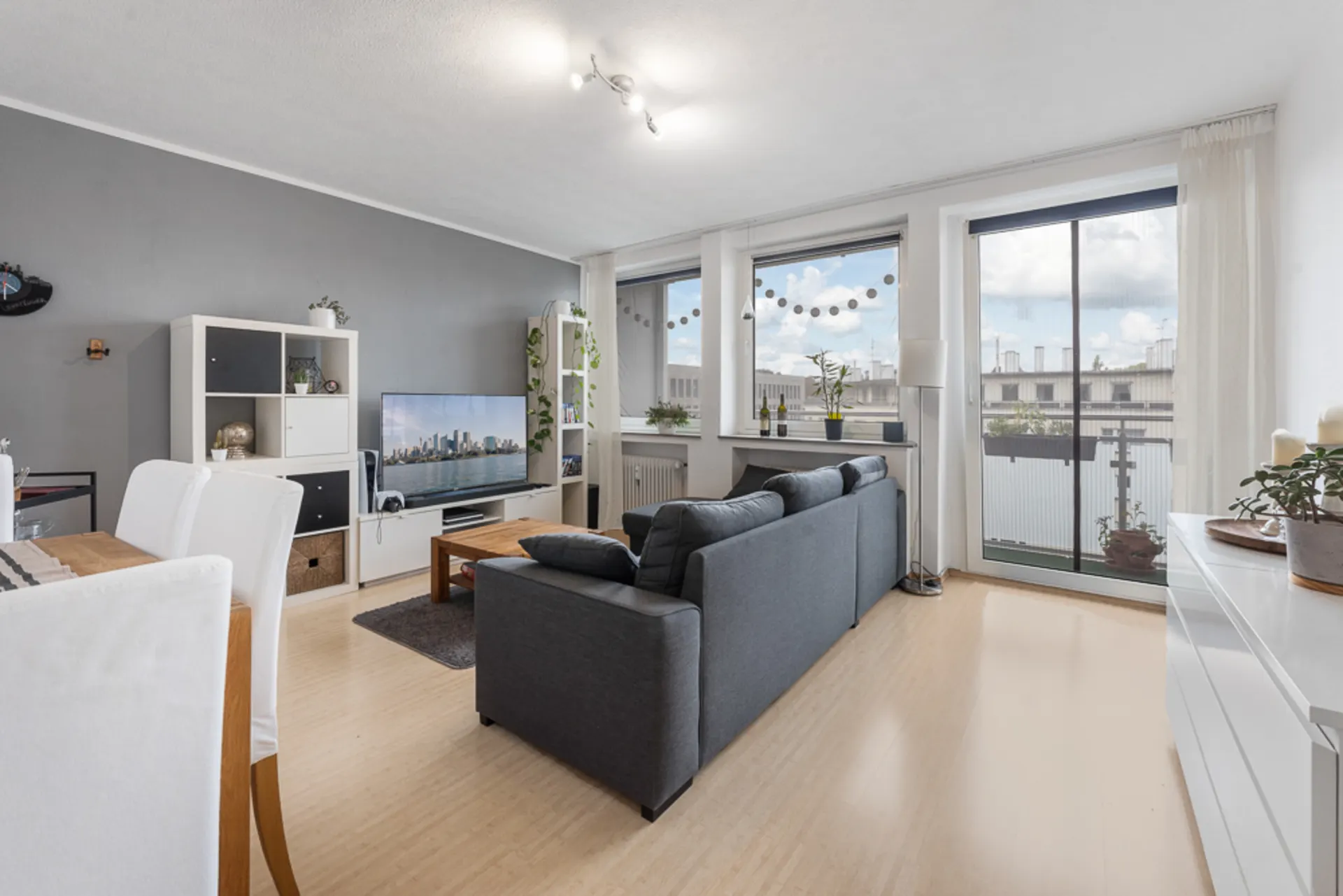 Helles Wohnzimmer mit grauer Couch, Fernseher, Esstisch und großen Fenstern mit Balkonzugang.