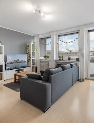 Helles Wohnzimmer mit grauer Couch, Fernseher, Esstisch und großen Fenstern mit Balkonzugang.