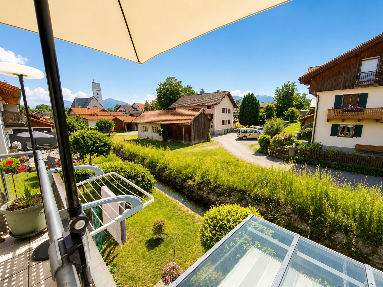 Blick von einem Balkon auf Gärten, einen kleinen Bach und Nachbarhäuser bei Sonnenschein.
