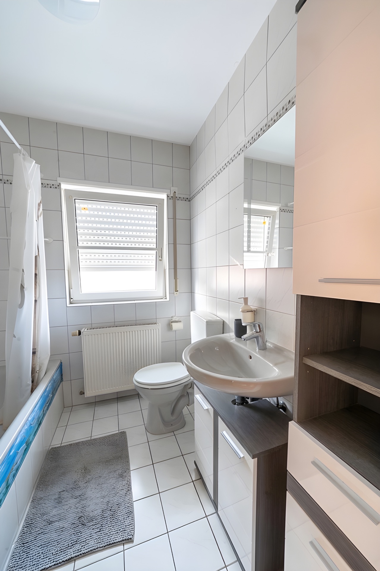 Helles Badezimmer mit Badewanne, WC, Waschbecken, Spiegel und Fenster mit Jalousie.