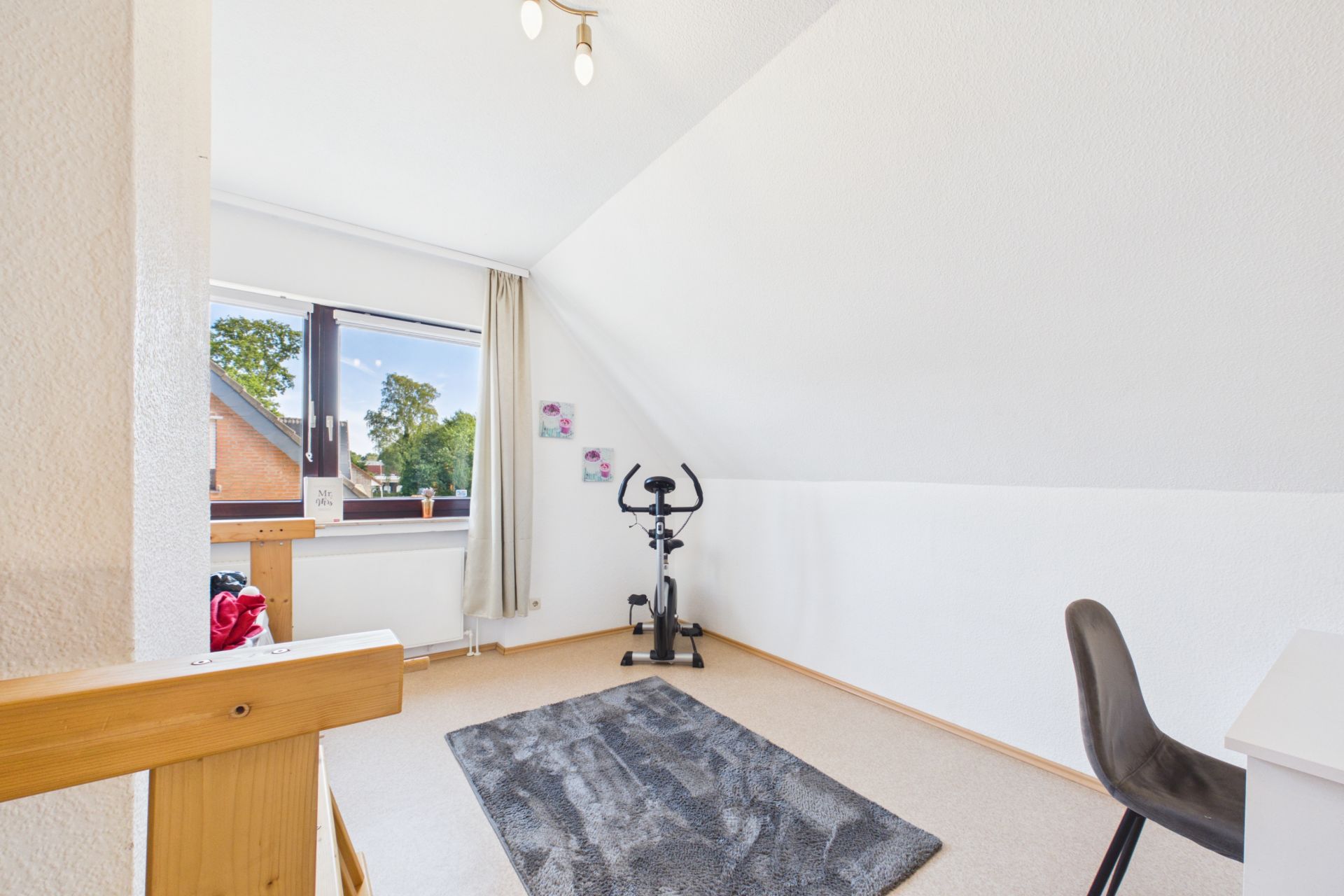 Helles Zimmer mit Schrägdach, Teppich, Heimtrainer, Schreibtischstuhl und Fenster.