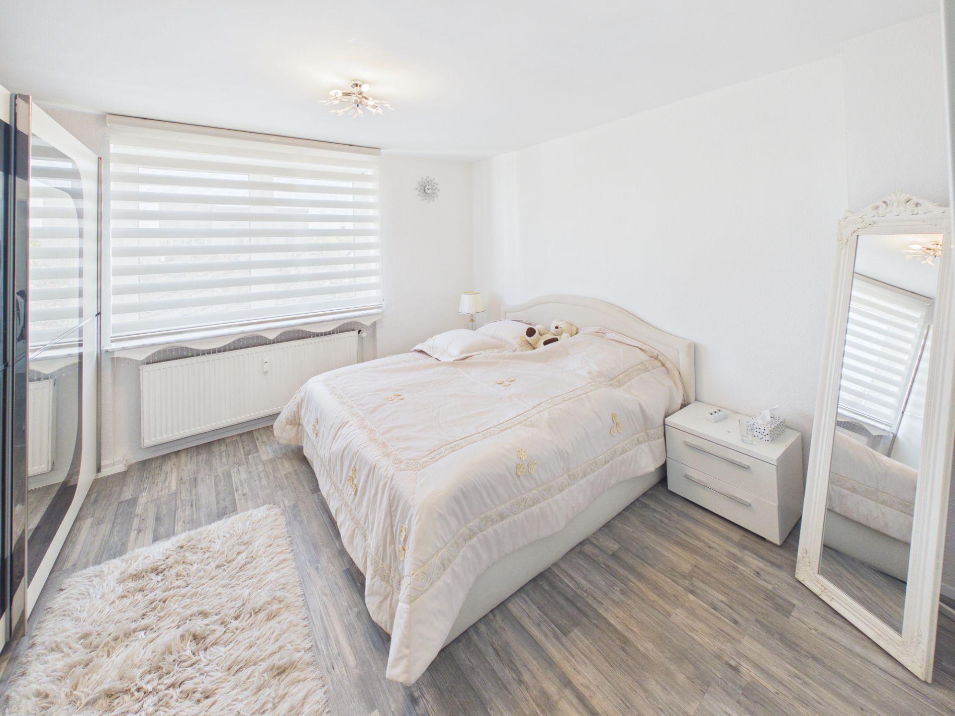 Helles Schlafzimmer mit großem Bett, Nachttisch, Spiegel und Fenster mit Jalousien.