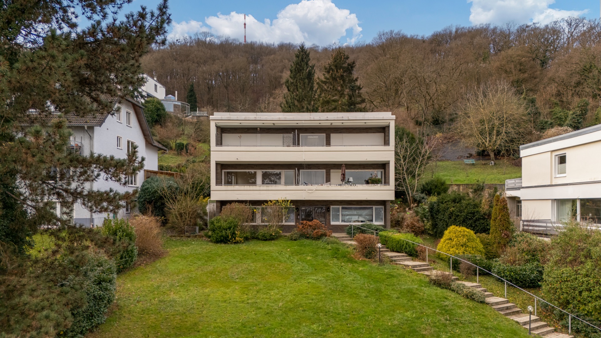 Dreistöckiges Wohnhaus mit Balkon, großer Garten und umliegenden Bäumen vor einem bewaldeten Hang.