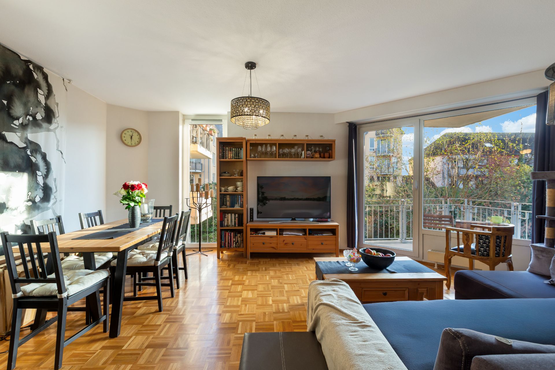 Helles Wohnzimmer mit Esstisch, Sofa, TV, Bücherregal und großen Fenstern mit Blick nach draußen.