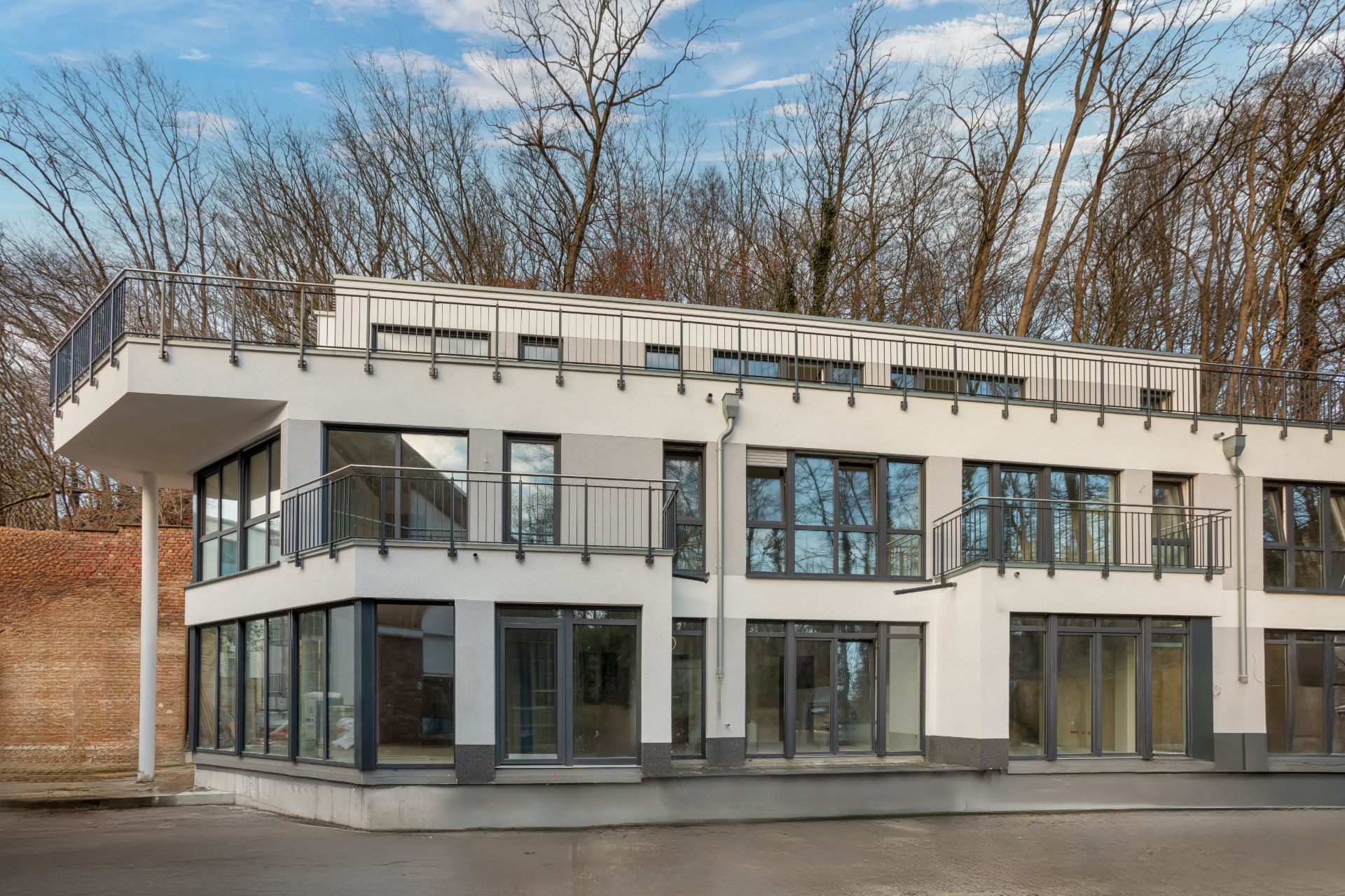 Modernes zweistöckiges Wohnhaus mit großen Fenstern und mehreren Balkonen, im Hintergrund Bäume.